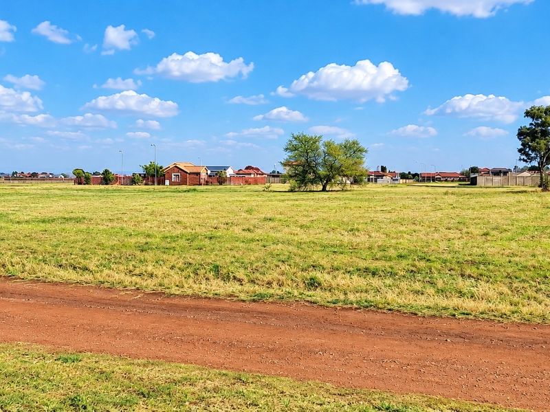 A wide, open residential vacant land plot featuring natural grassland and a dirt access path under a clear blue sky.