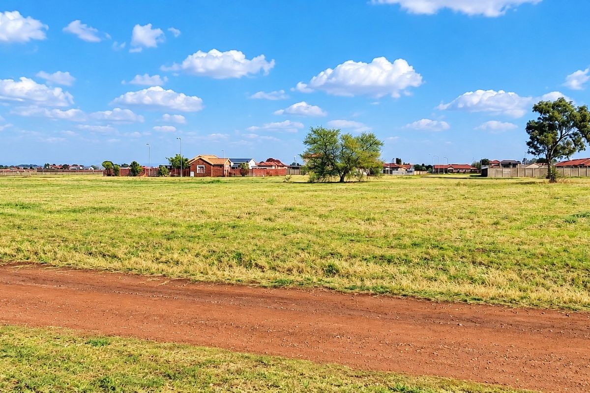A wide, open residential vacant land plot featuring natural grassland and a dirt access path under a clear blue sky.