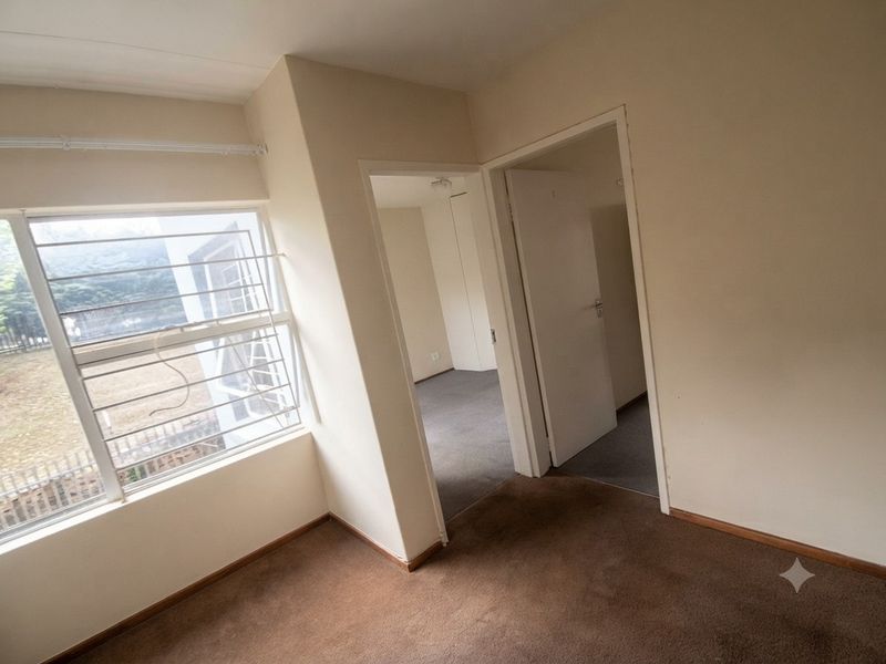 An empty bedroom featuring carpeted flooring, security bars on the window, and access to adjacent rooms through internal doorways.