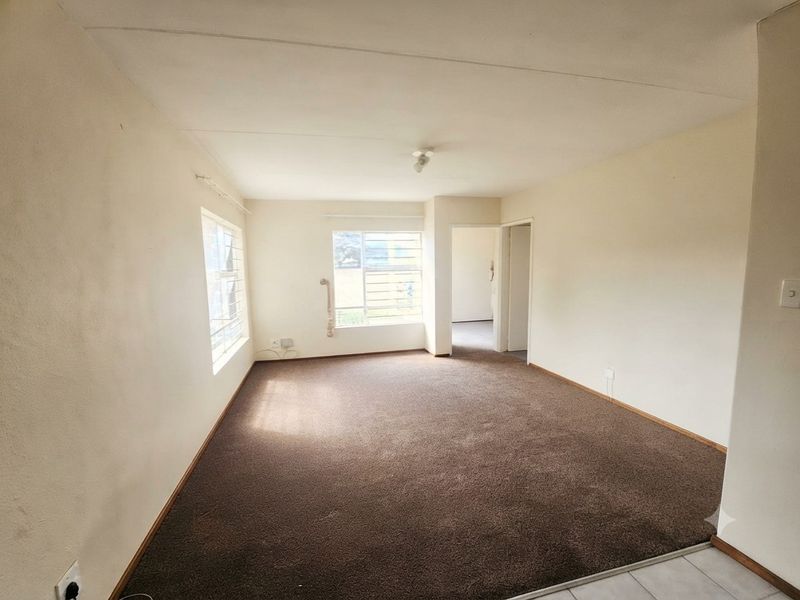 An empty living room featuring brown carpeting, neutral walls, and windows fitted with security burglar bars.