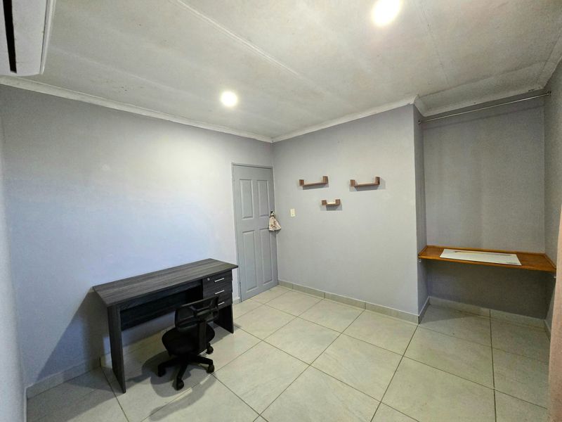 A functional bedroom featuring tiled flooring, a built-in desk area, wall-mounted shelving, and an air conditioning unit.