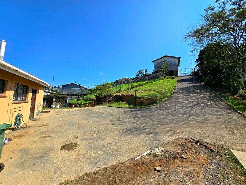 A wide-angle view of the property exterior featuring a large asphalt driveway, sloped garden area, and access to the main building structure.