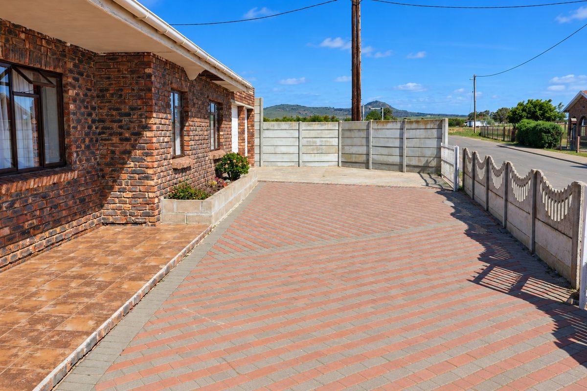 A neat front exterior featuring a face brick facade, a tiled patio, and a paved driveway enclosed by precast concrete boundary walls.