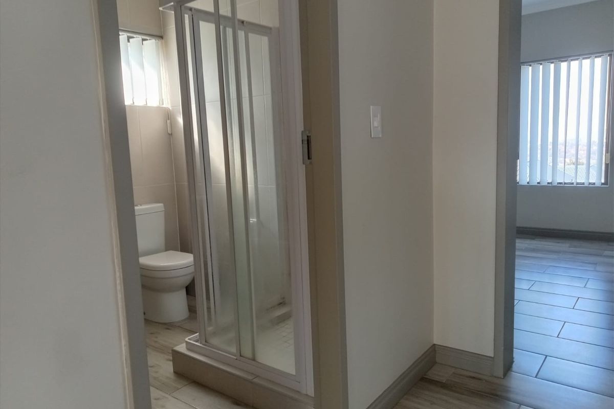 A functional bathroom featuring a shower enclosure and toilet, with neutral tiled flooring and vertical blinds.