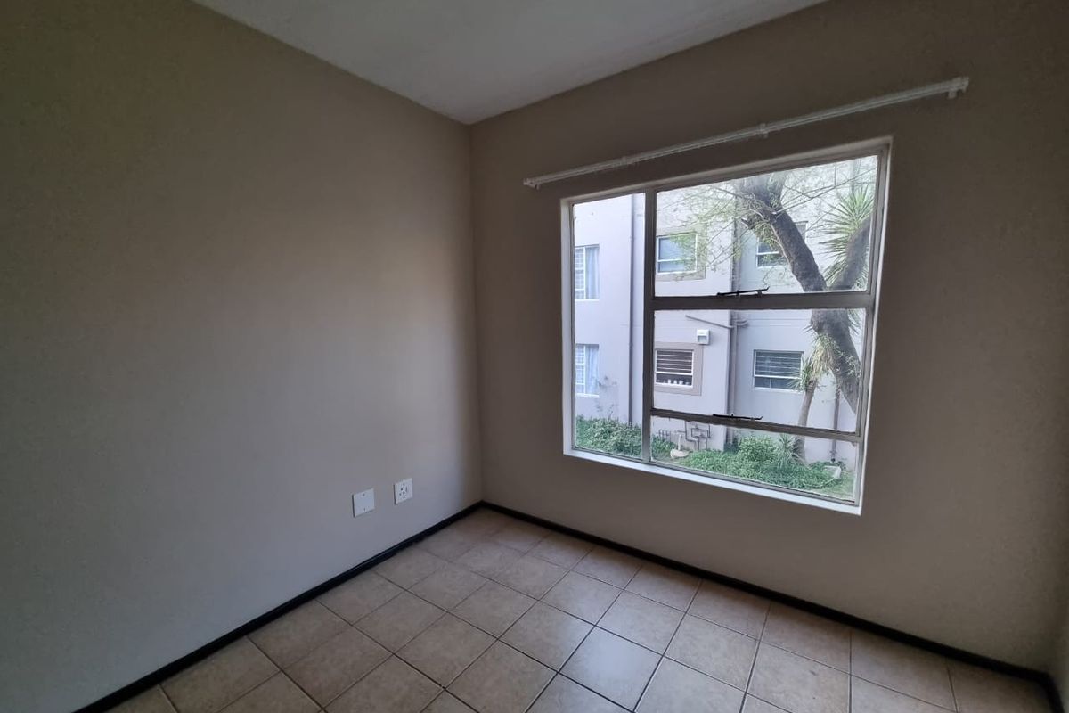 A clean, empty bedroom featuring neutral wall paint, tiled flooring, and a large window providing natural light and views of the complex exterior.