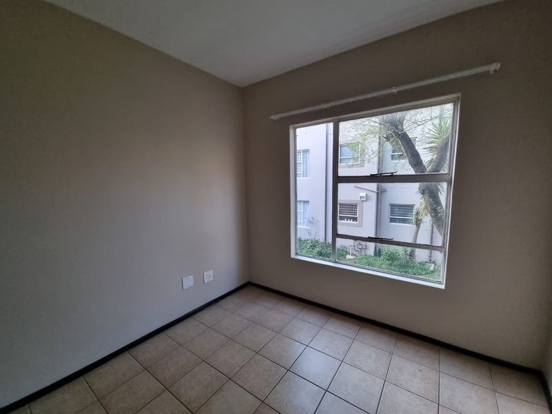 A clean, empty bedroom featuring neutral wall paint, tiled flooring, and a large window providing natural light and views of the complex exterior.