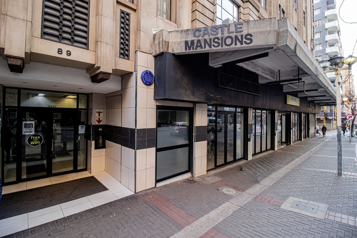 Street-level view of the Castle Mansions commercial building entrance, featuring a prominent canopy, glass storefronts, and tiled exterior facade.
