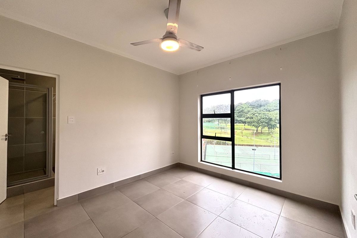 A bright, empty bedroom featuring neutral tiled flooring, a modern ceiling fan, and a large window with views of the surrounding estate.