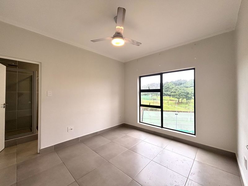 A bright, empty bedroom featuring neutral tiled flooring, a modern ceiling fan, and a large window with views of the surrounding estate.