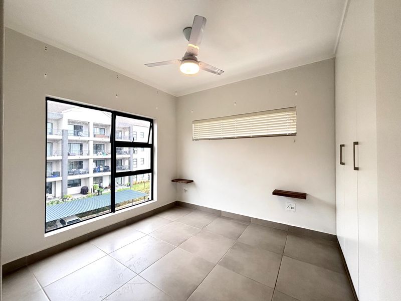 A bright, empty bedroom featuring neutral tiled flooring, built-in cupboards, a modern ceiling fan, and large aluminium-framed windows with blinds.