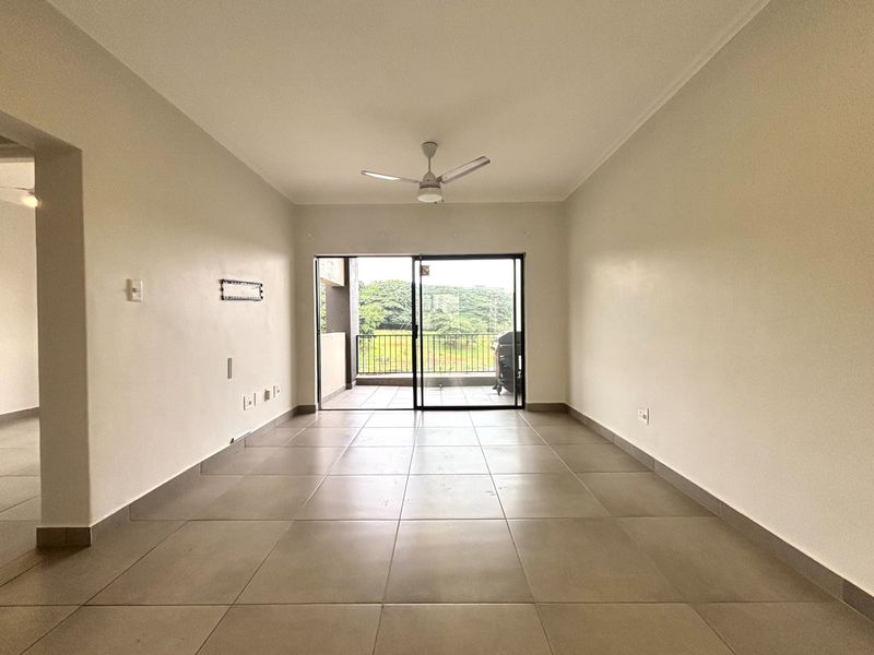 An empty, well-lit living room featuring neutral tiled flooring, a ceiling fan, and sliding glass doors leading to an outdoor balcony area.