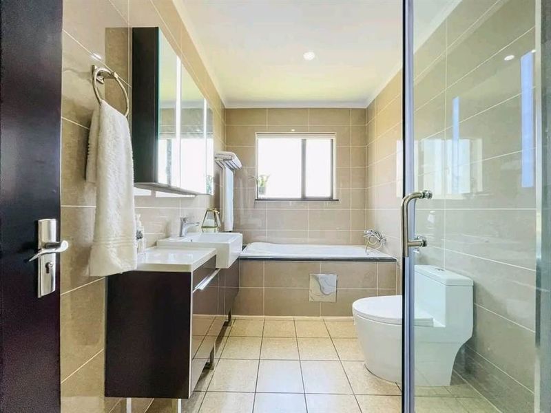 A modern, well-maintained bathroom featuring a built-in vanity, bathtub, glass-enclosed shower, and neutral floor-to-ceiling tiling.