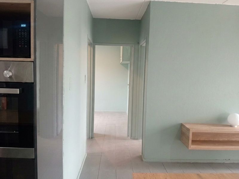 A bright hallway featuring light green walls, timber flooring, and integrated kitchen appliances, leading into an adjacent room.