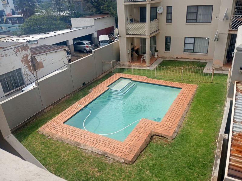 Aerial view of a communal apartment complex pool area featuring a brick-edged swimming pool, surrounding lawn, and metal fencing.
