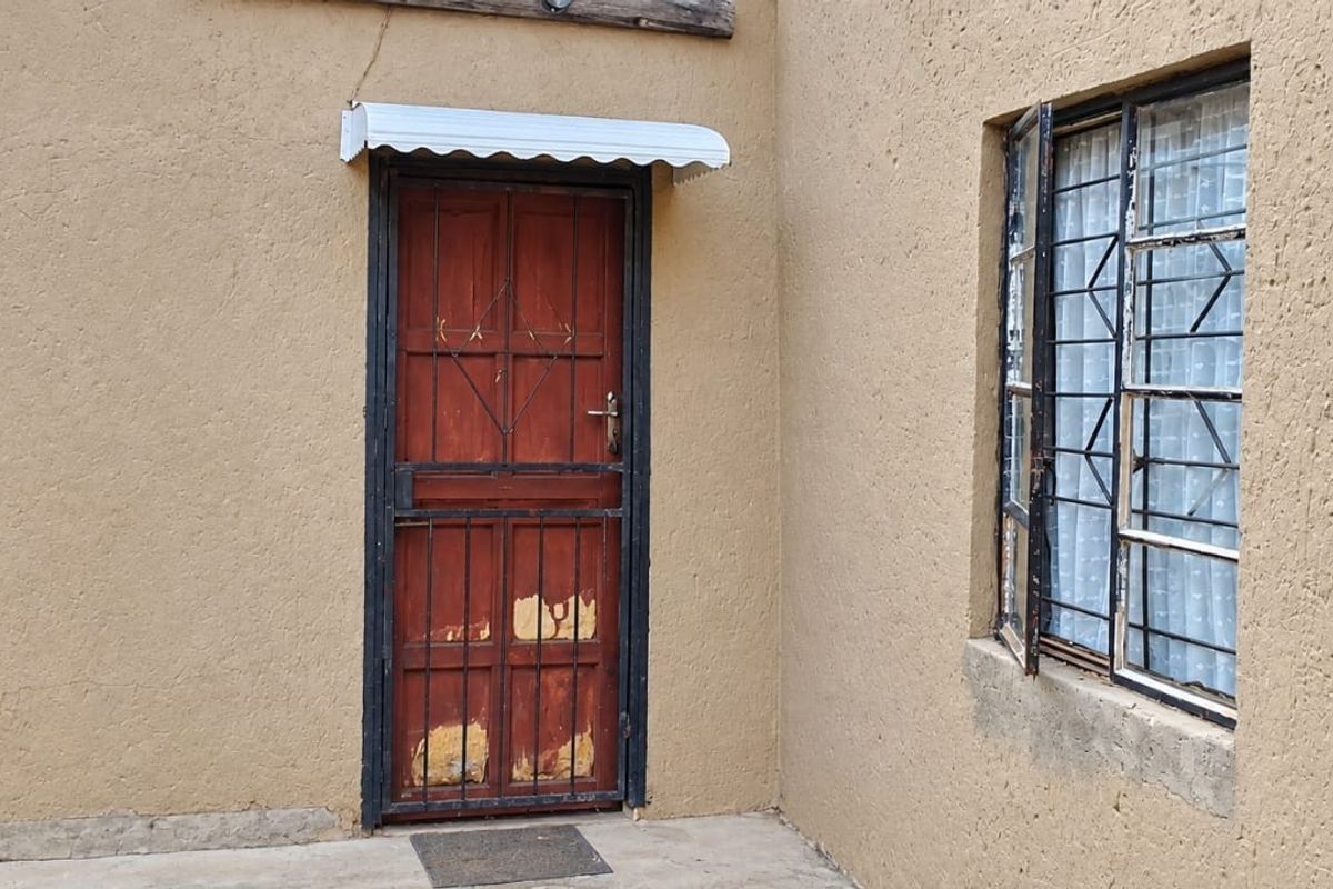 Rear exterior view showing a textured wall finish, a wooden door with a security gate, and protective window security bars.