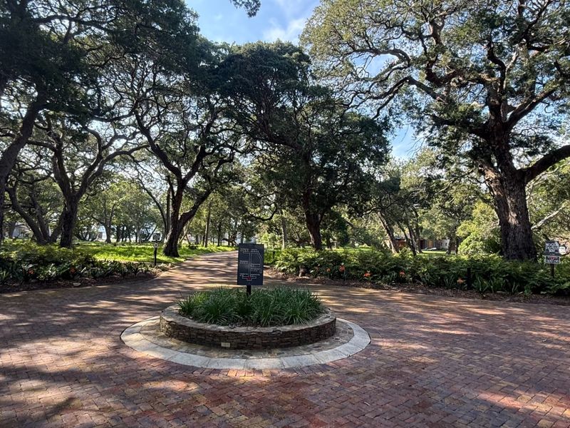 A beautifully landscaped entrance area featuring a circular brick-paved driveway, a stone-walled central planter, and mature trees providing a serene, shaded environment.