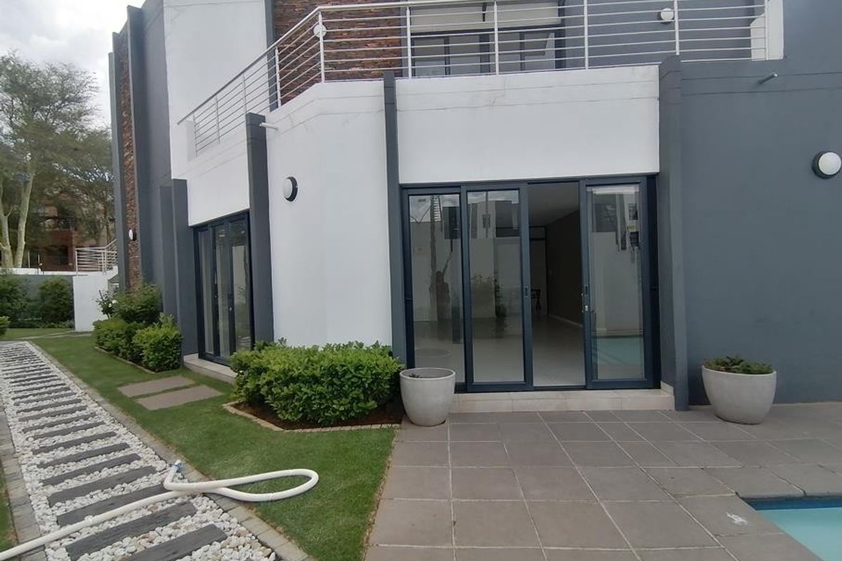 Rear view of a modern apartment featuring a tiled patio, swimming pool, landscaped garden, and a balcony with stainless steel railings.