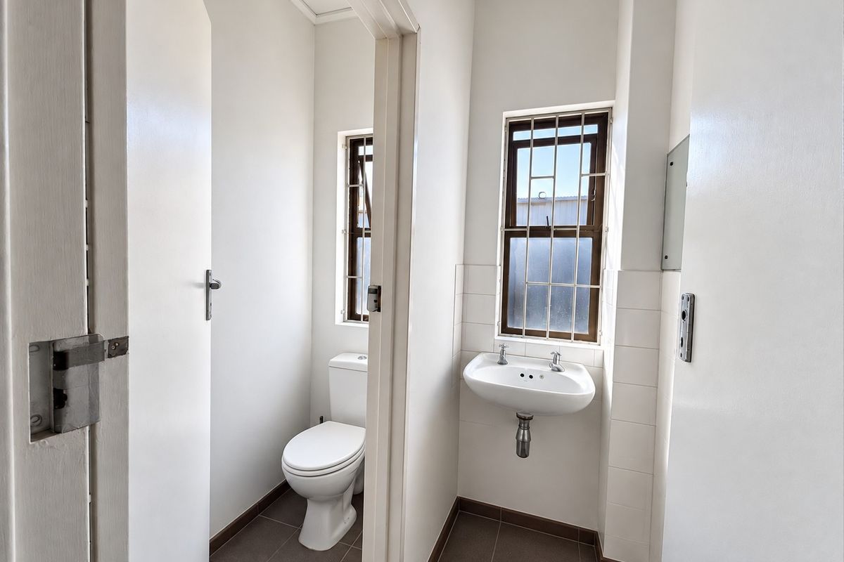 A functional industrial-style bathroom featuring a separate toilet cubicle and a wall-mounted basin with tiled flooring and security-barred windows.