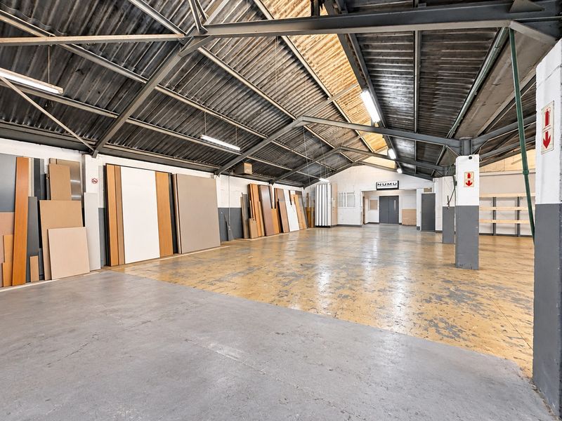 Spacious industrial warehouse unit featuring high exposed steel roof trusses, concrete flooring, and functional strip lighting, suitable for various commercial or storage applications.