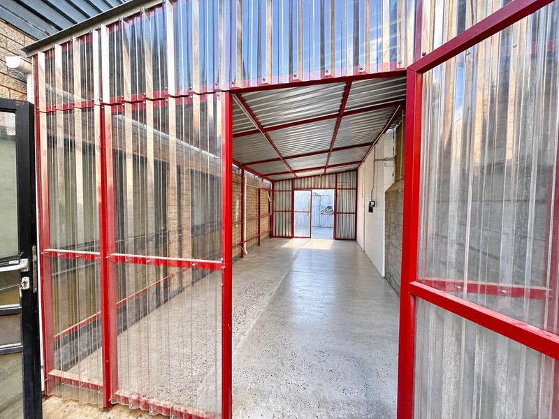 A covered industrial utility walkway or storage area featuring a red steel frame, corrugated polycarbonate walls, and durable concrete flooring.