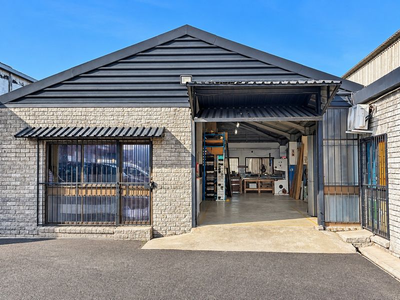 Front elevation of an industrial unit featuring a brick facade, security bars on windows, and a large roller shutter door providing access to the workspace.