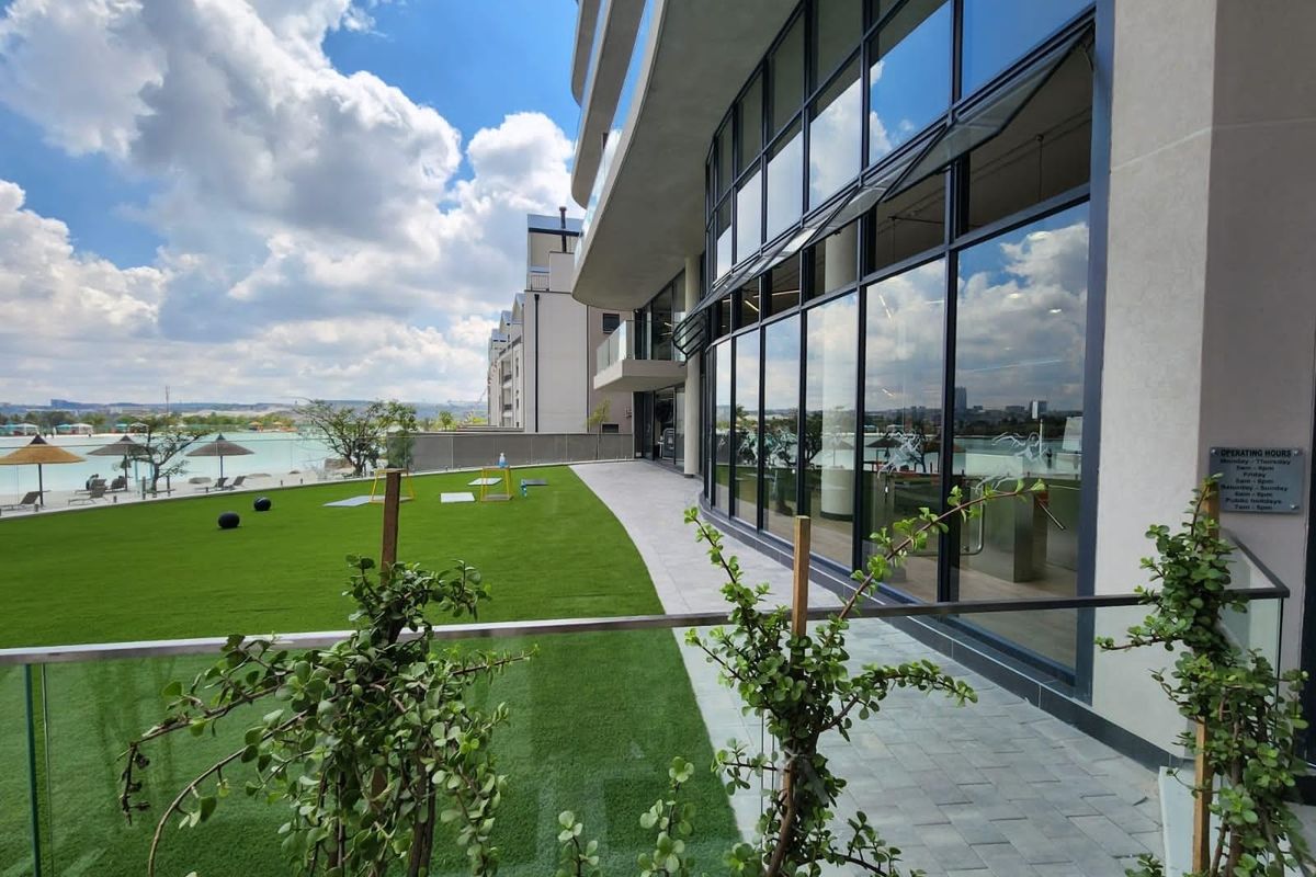 Modern apartment exterior featuring a glass facade, paved walkway, and a well-maintained artificial lawn overlooking a lagoon area.