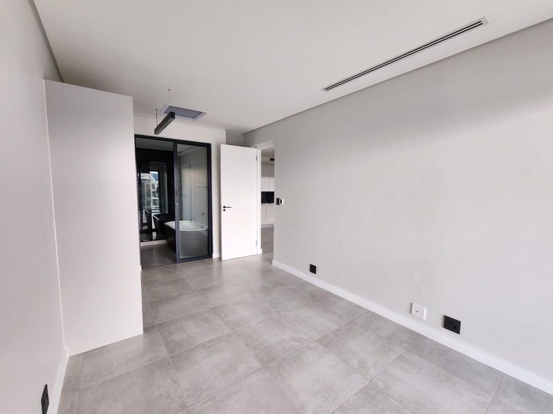 A modern, empty bedroom featuring neutral tiled flooring, ducted air conditioning, and a sleek glass partition leading to an en-suite bathroom.