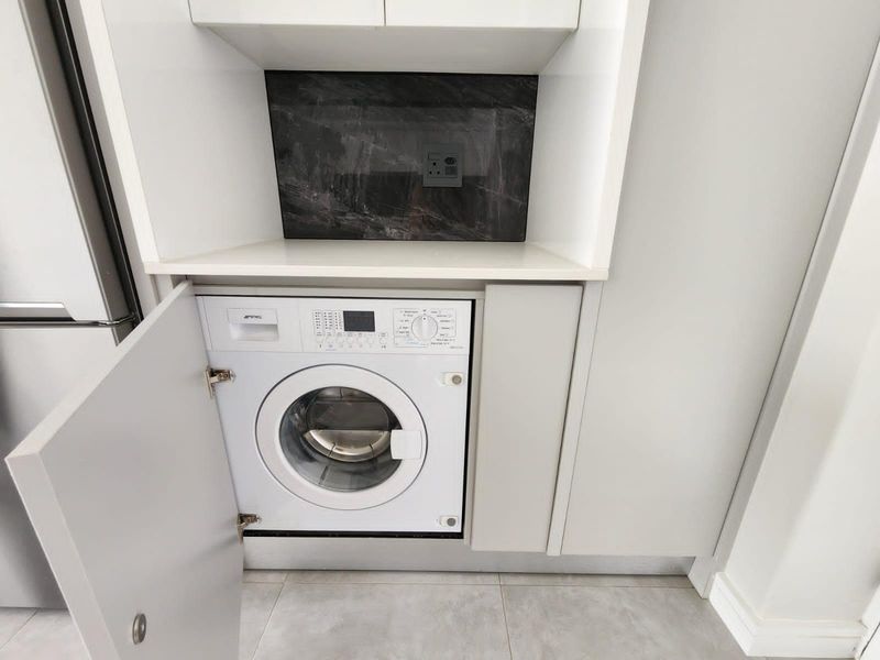 A compact, modern laundry space featuring integrated cabinetry, a stone splashback, and a built-in washing machine with tiled flooring.