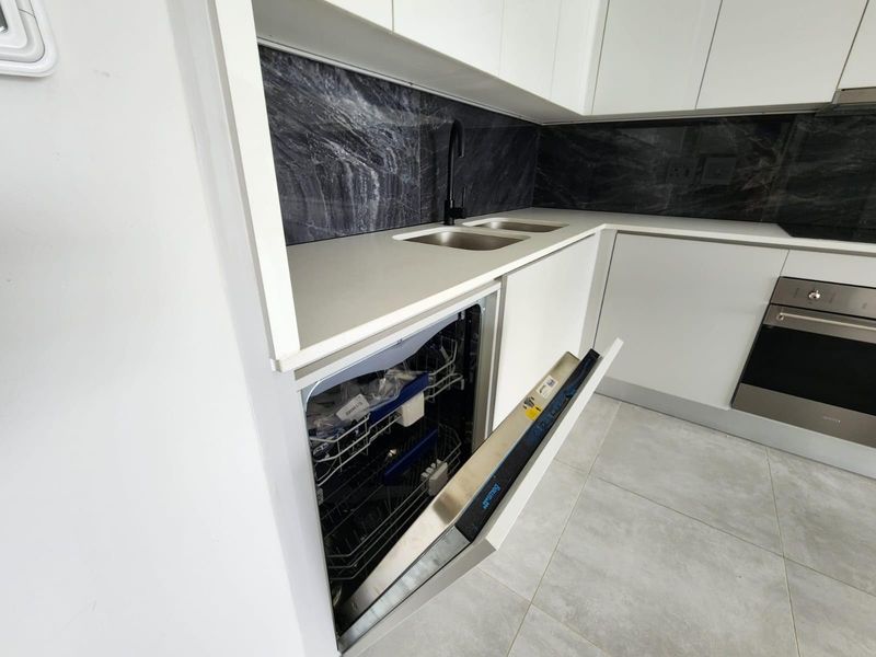 Modern kitchen featuring sleek handleless white cabinetry, stone countertops, a marble-look splashback, integrated dishwasher, and a built-in oven.