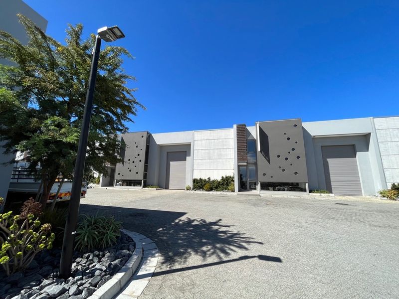 Modern industrial unit exterior featuring clean architectural lines, roller shutter doors, and paved driveway access in a well-maintained business park setting.