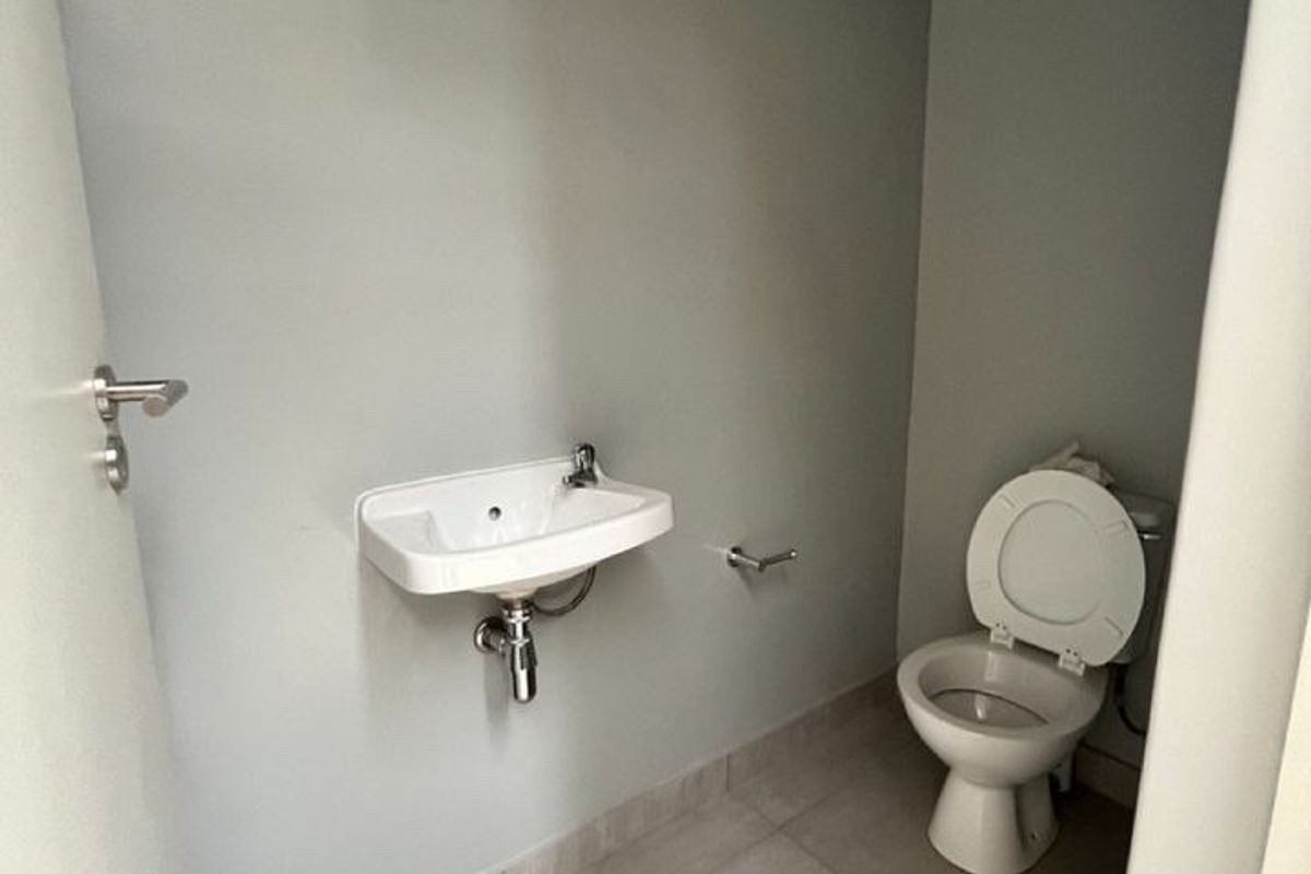 A compact industrial-style restroom featuring a wall-mounted basin, toilet, and exposed ceiling ducting with neutral tiled flooring.
