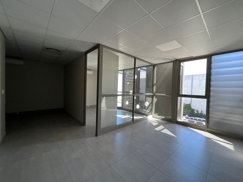 A bright, empty office space featuring modern glass partitioning, tiled flooring, and a suspended ceiling, suitable for a professional workspace.