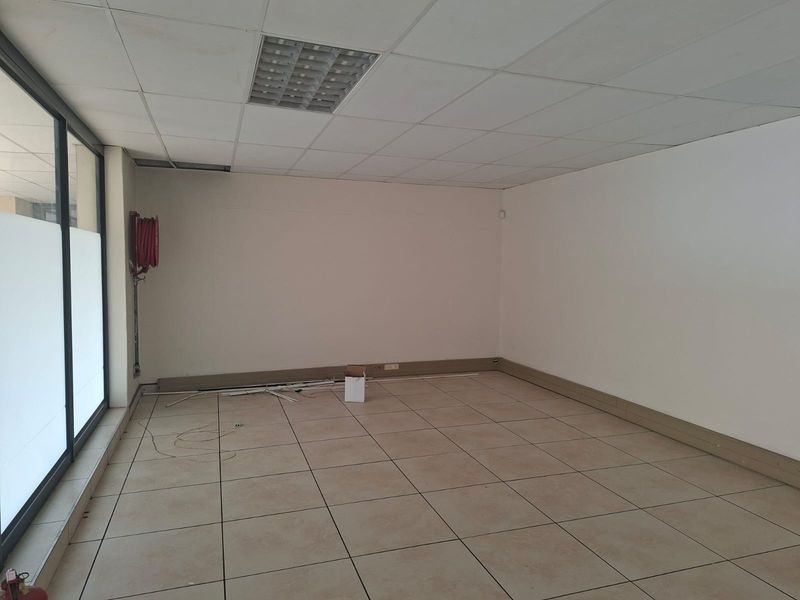 An empty office space featuring tiled flooring, a suspended ceiling with recessed lighting, and large windows providing natural light.
