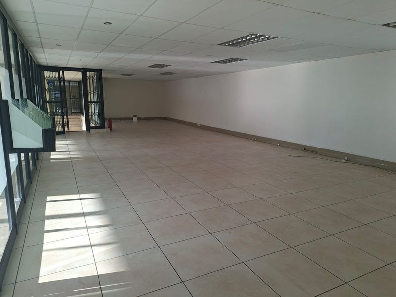 A spacious, vacant open-plan office area featuring tiled flooring, a suspended ceiling with recessed lighting, and large glass windows providing natural light.