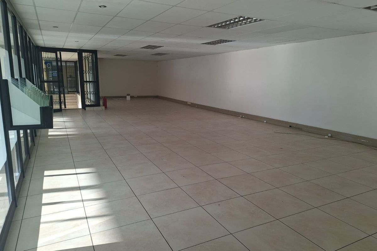 A spacious, vacant open-plan office area featuring tiled flooring, a suspended ceiling with recessed lighting, and large glass windows providing natural light.