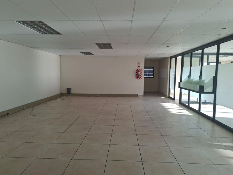 A spacious, empty office area featuring tiled flooring, a suspended ceiling with recessed lighting, and large glass partitions allowing for natural light.