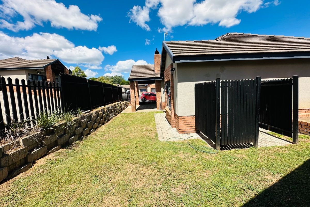 Rear exterior view of the property featuring a lawn, paved walkway, brick retaining wall, and secure perimeter fencing.