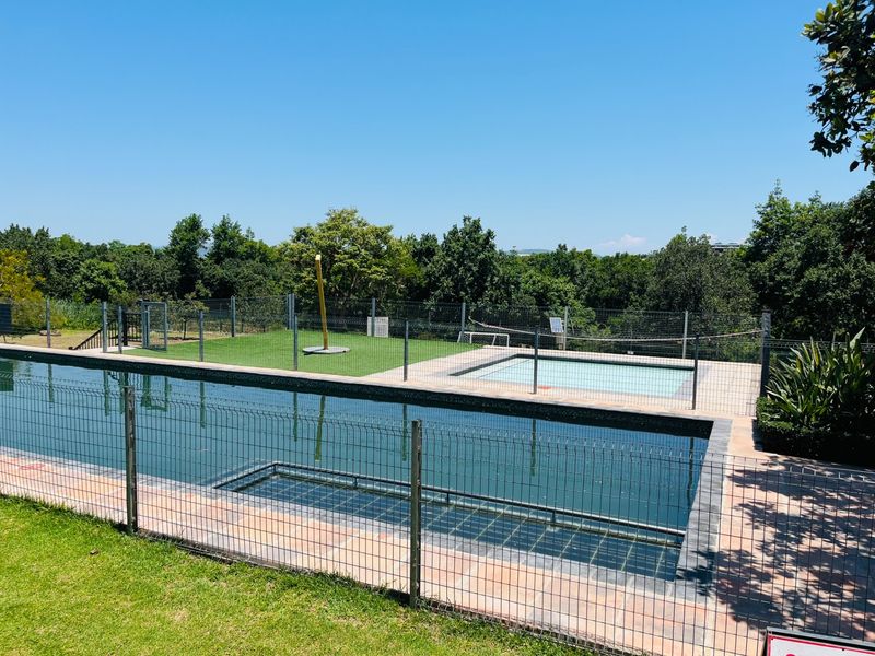 A well-maintained outdoor pool area featuring a large swimming pool, a separate wading pool, safety fencing, and a paved surround with artificial grass.