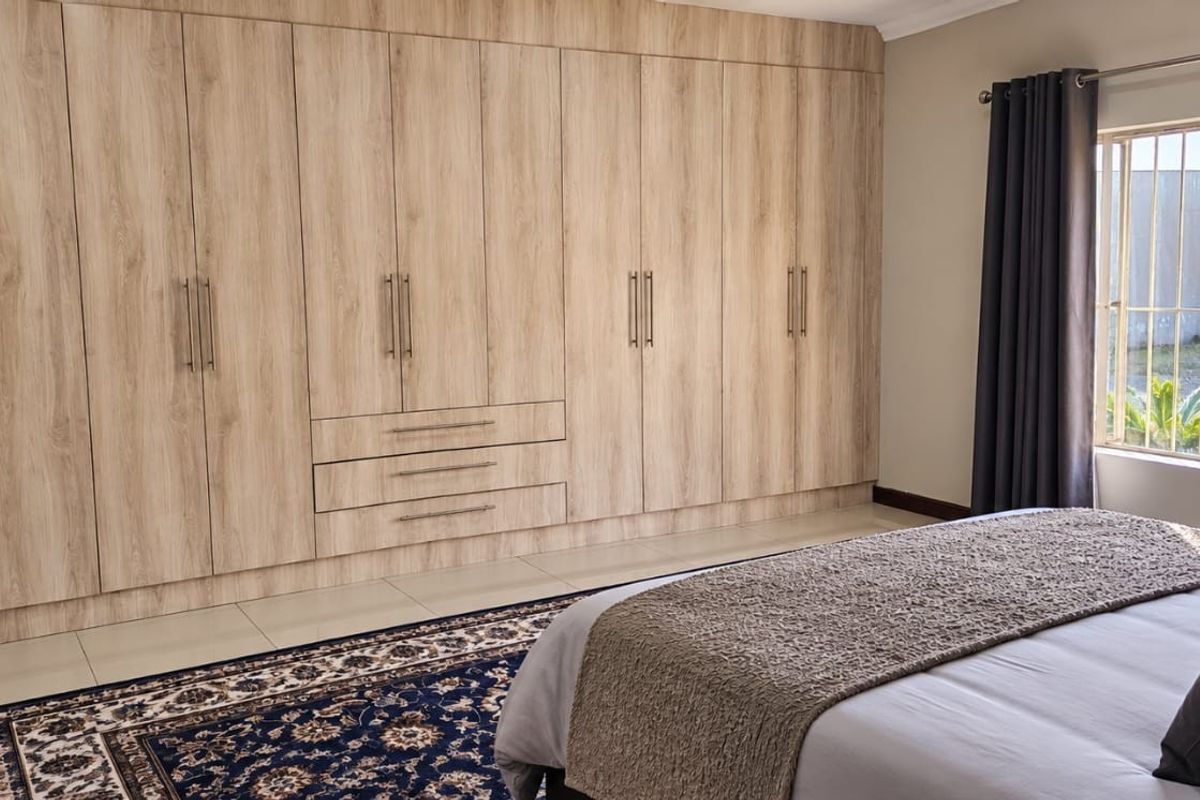 A well-maintained bedroom featuring extensive light wood-grain built-in cupboards, tiled flooring, and large windows fitted with security bars.