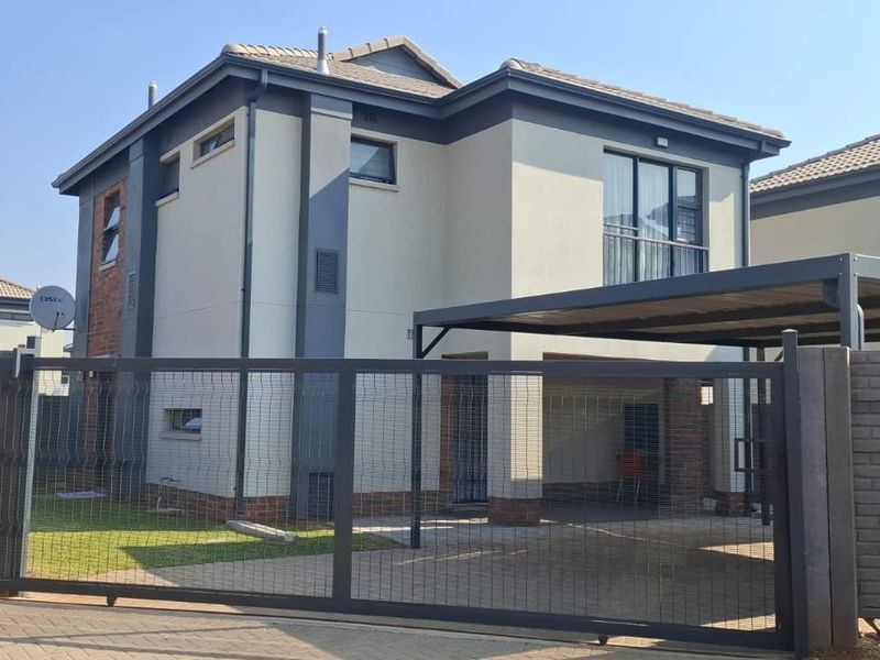 A modern double-storey residential property featuring a paved driveway, secure sliding gate, carport, and a balcony, set within a neat brick-walled perimeter.