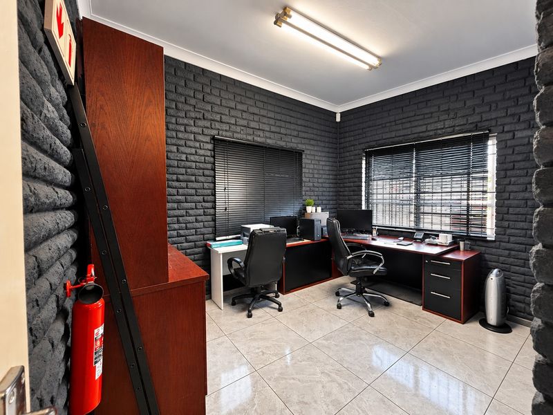A functional home office featuring dark painted exposed brick walls, neutral tiled flooring, and dual windows with venetian blinds for natural light control.