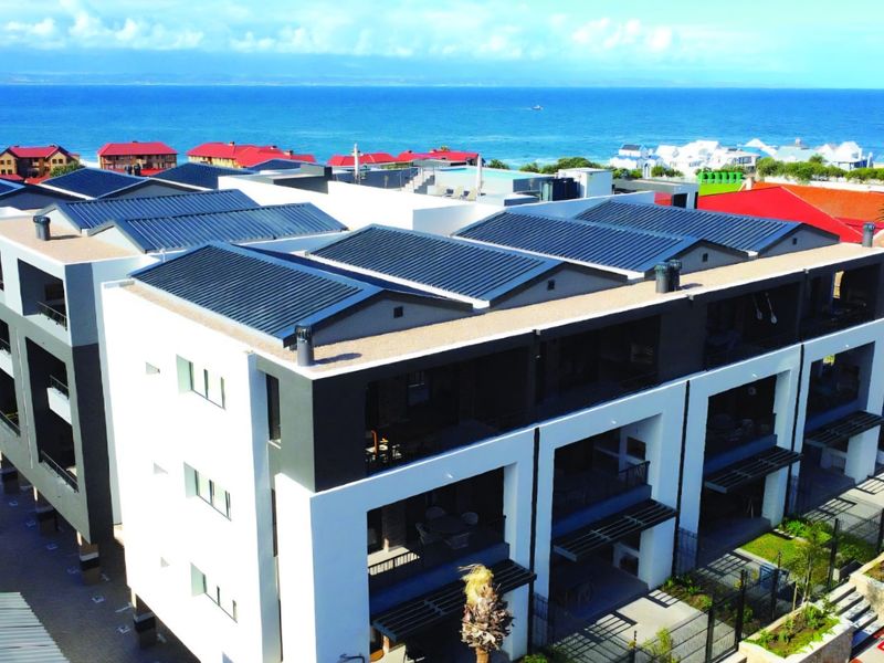 Aerial view of a modern apartment complex featuring metal roofing, private balconies, and landscaped grounds with a scenic ocean backdrop.