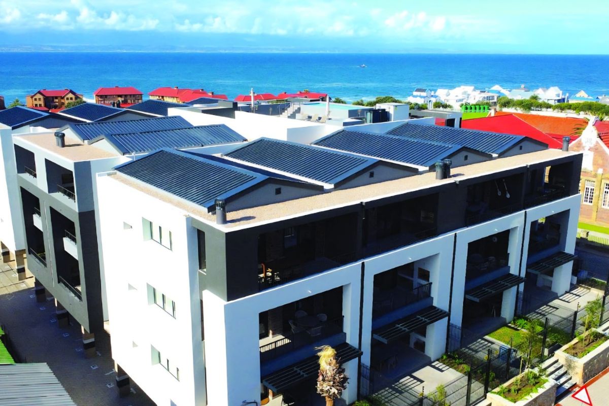 Aerial view of a modern apartment complex featuring metal roofing, private balconies, and landscaped grounds with a scenic ocean backdrop.