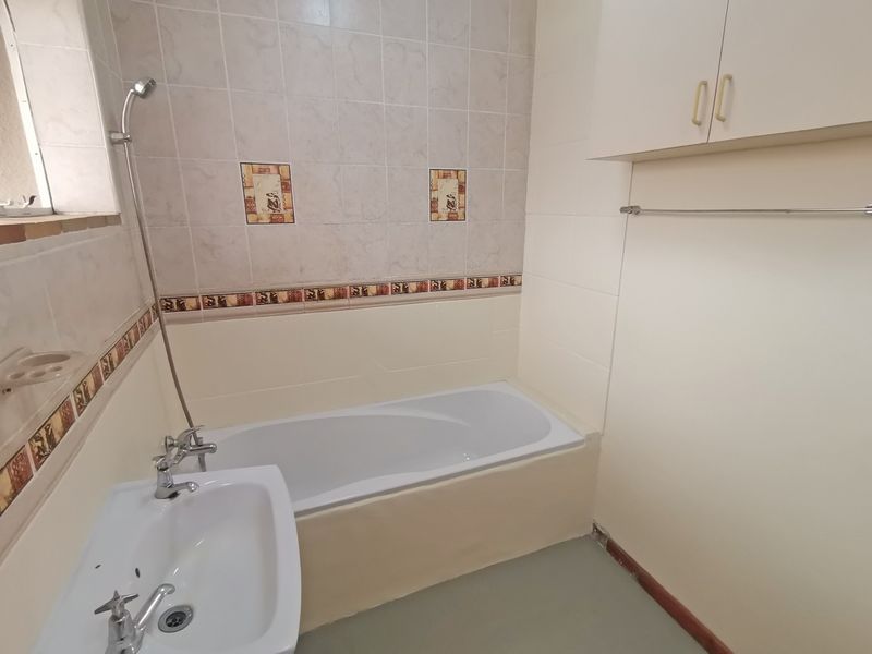 A functional bathroom featuring a built-in bathtub, wall-mounted shower, pedestal basin, and overhead storage cabinetry with neutral wall tiling.