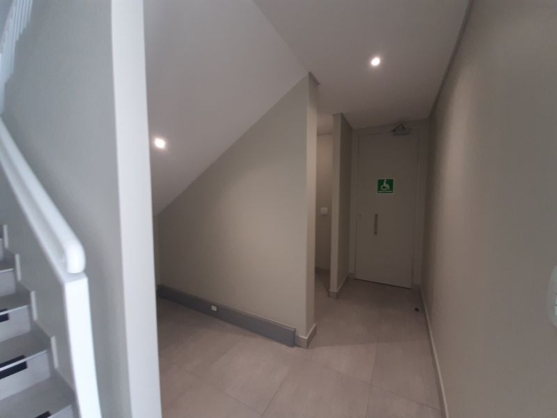 A clean, modern hallway featuring tiled flooring, recessed lighting, and an accessible doorway, with an adjacent staircase.