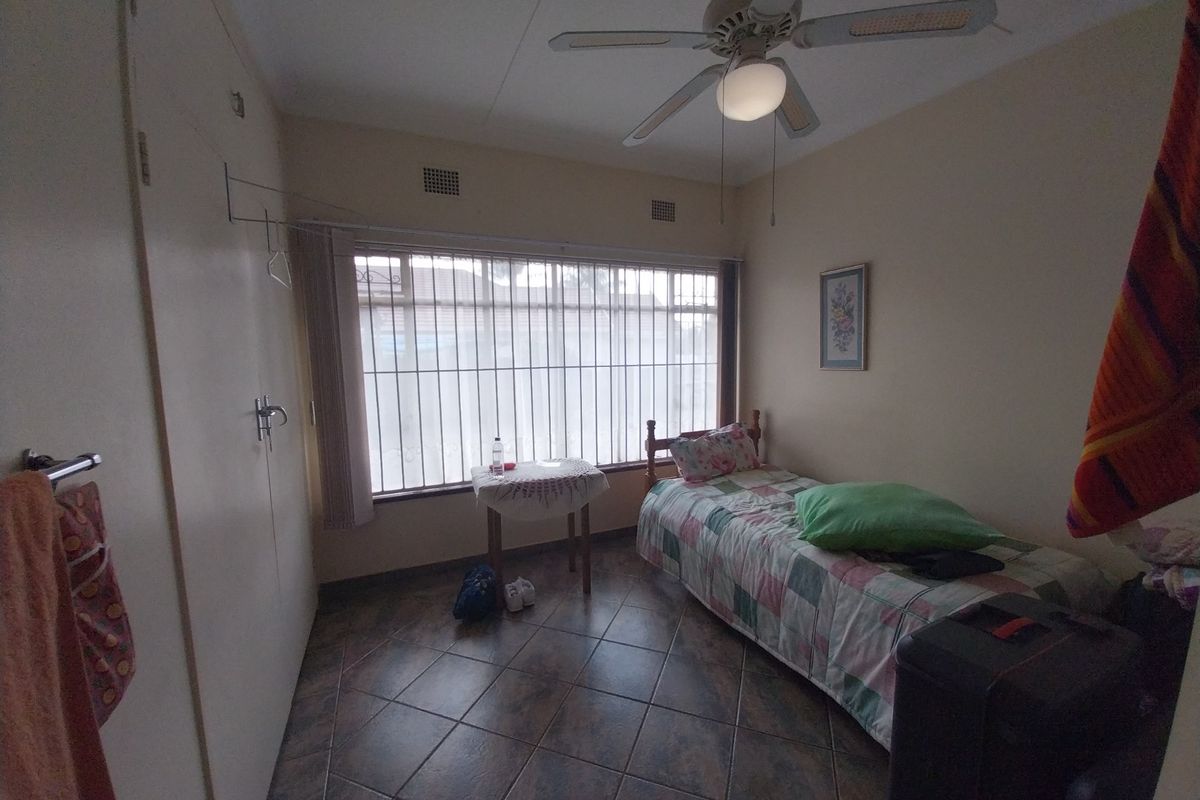 A functional bedroom featuring tiled flooring, built-in cupboards, and a ceiling fan, with large windows secured by metal bars.