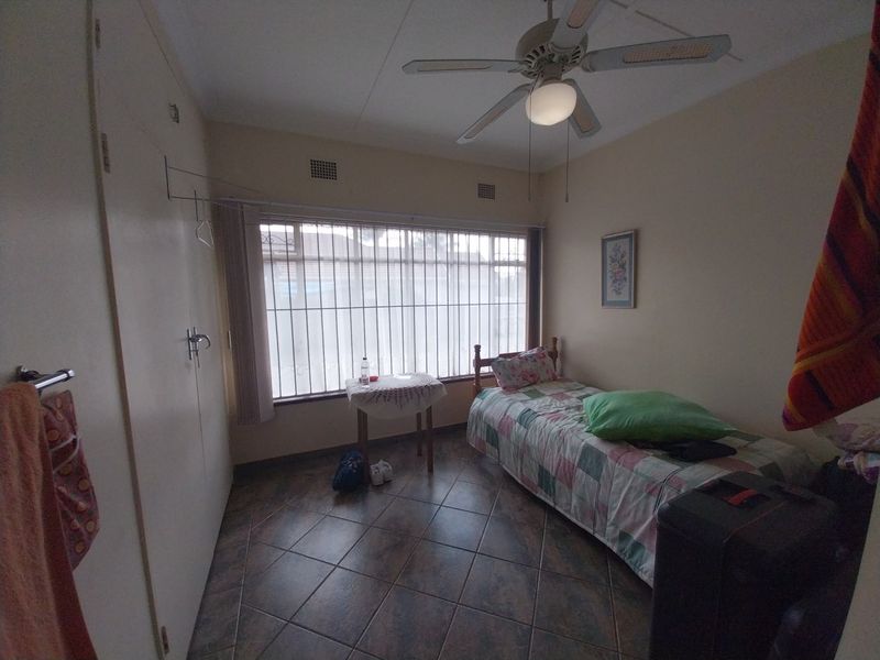 A functional bedroom featuring tiled flooring, built-in cupboards, and a ceiling fan, with large windows secured by metal bars.