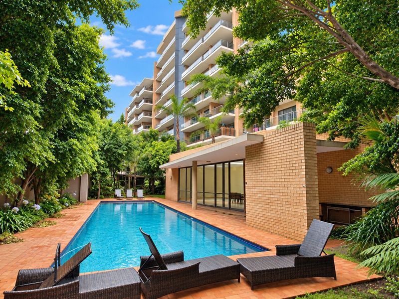 A well-maintained outdoor swimming pool area featuring a brick facade, paved deck, and direct access to the apartment building through sliding glass doors.