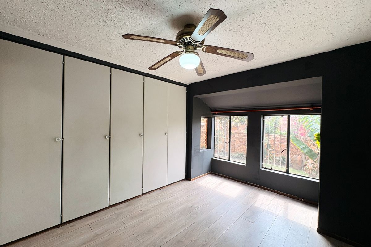 A spacious bedroom featuring extensive built-in cupboards, laminate flooring, and a ceiling fan, with large windows providing natural light and garden views.