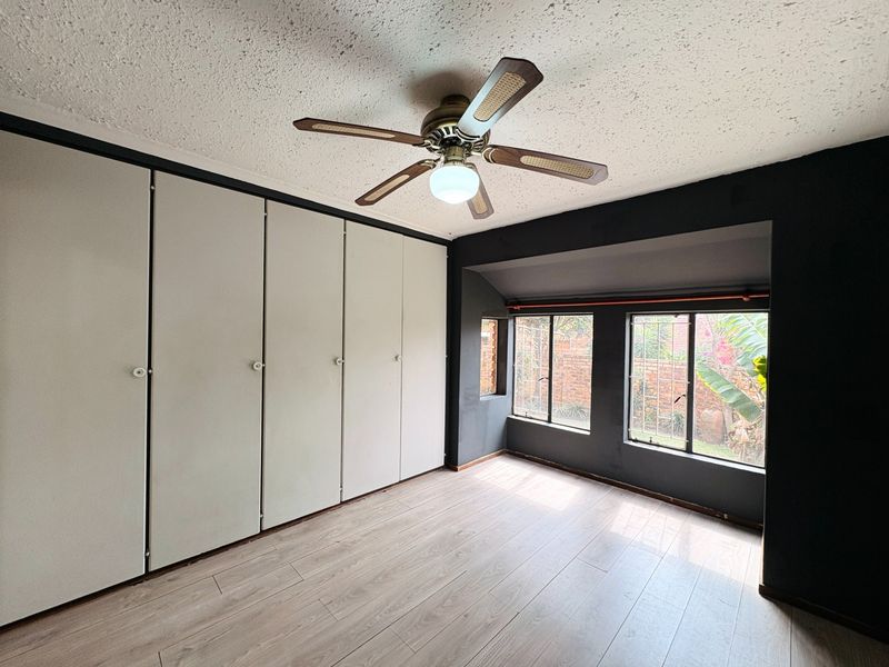 A spacious bedroom featuring extensive built-in cupboards, laminate flooring, and a ceiling fan, with large windows providing natural light and garden views.